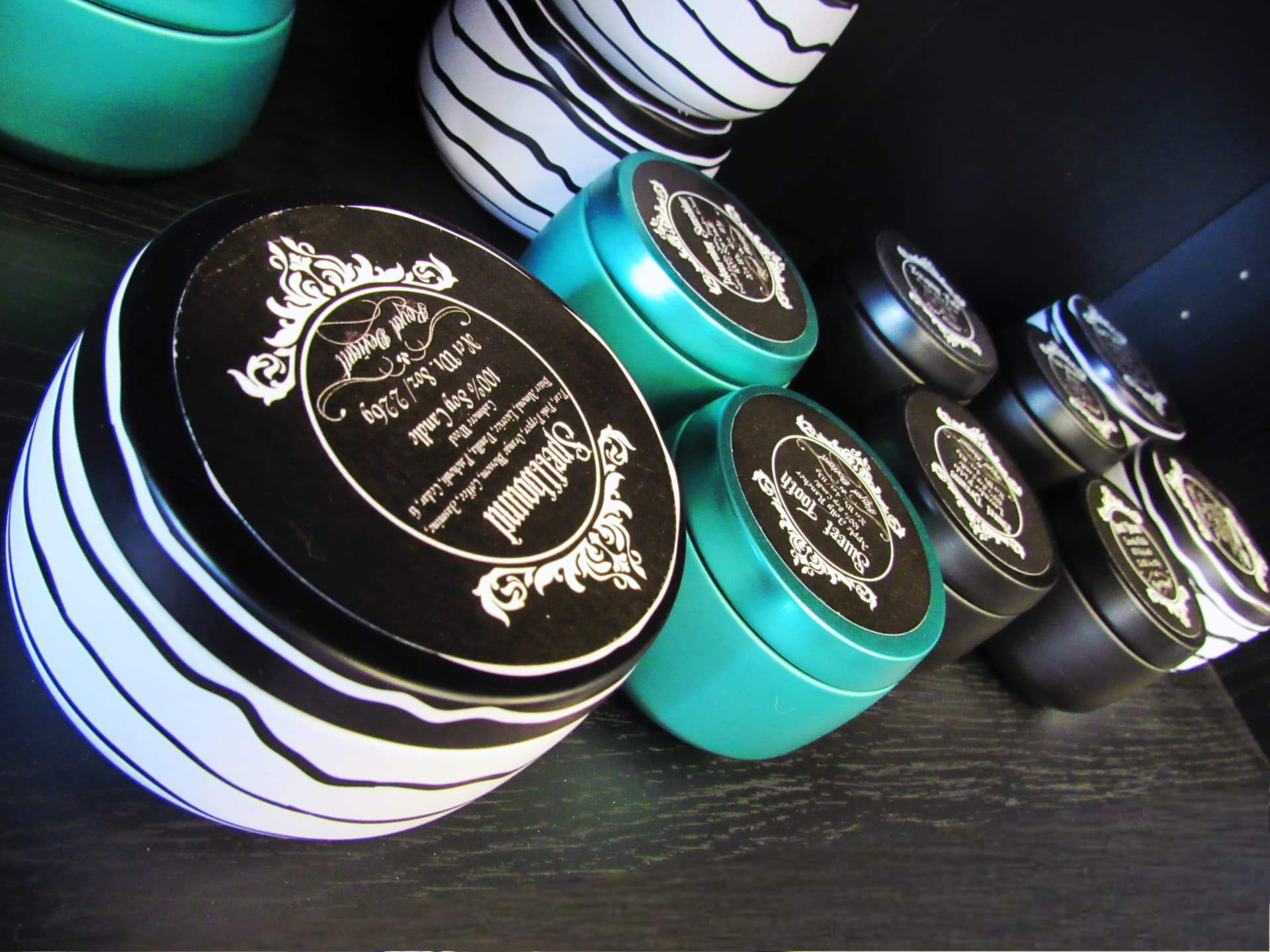 Black and teal candle tins with ornate labels stacked on a dark shelf